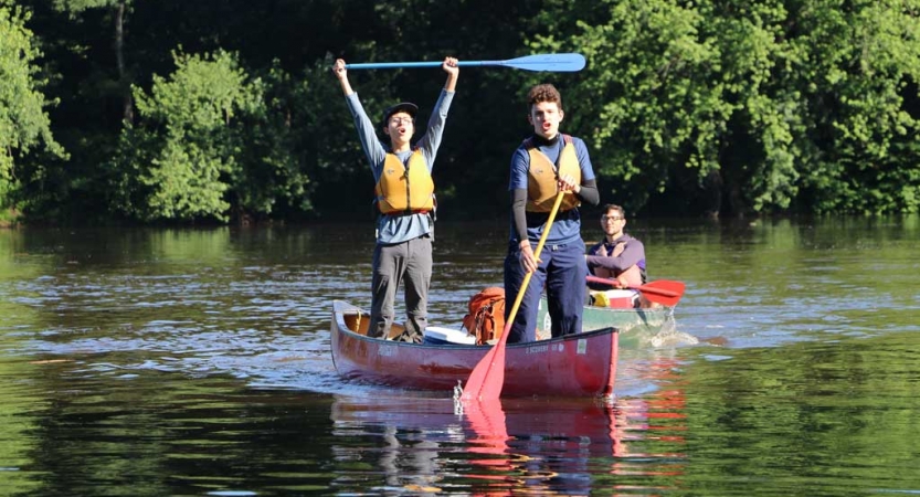 Two people wearing lifejackets stand in a canoe floating on water. 